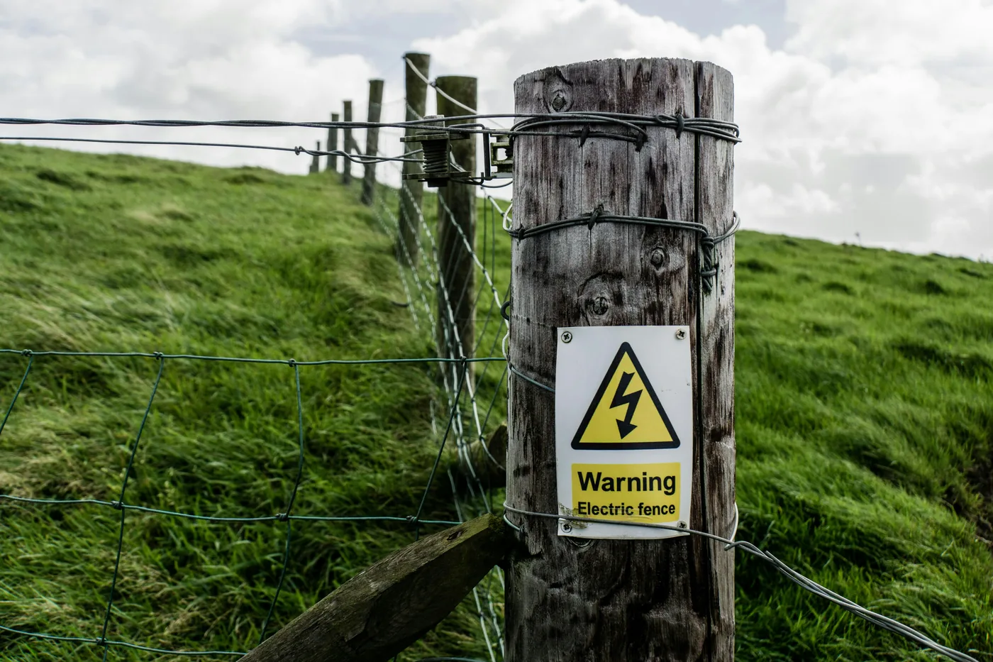 Wooden fence post with an electric fence warning sign on a grassy pasture